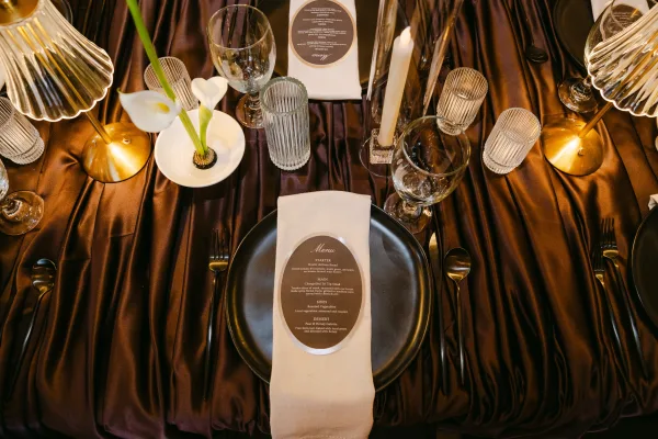 Reception tablescape with a modern black place setting on satin table linen, warmed by the glow of brass table lamps.
