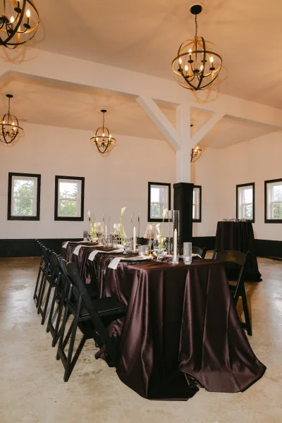 Reception tablescape styled on a chocolate satin tablecloth with calla lilies, arranged for a modern indoor wedding dinner along a long banquet table