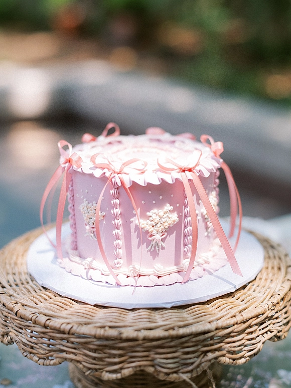 Wedding cake with pink wedding cake ribbon bows, scalloped piping, pearl sprinkles and heart details on a wicker stand in a blurred garden backdrop