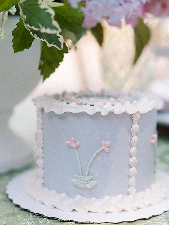 Wedding cake with pastel frosting, piped ruffle icing and floral detail, topped with a heart topper on a greenery-lined board at a blurred table setting