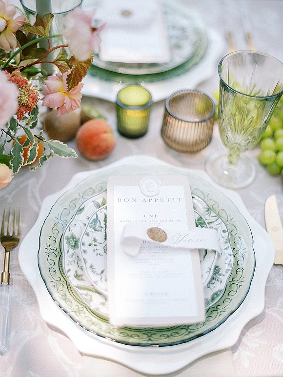 Reception tablescape with wedding place setting featuring layered floral china, pink centerpiece, green goblet, gold flatware, and candles on white linen