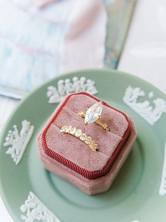 Wedding rings featuring a marquise engagement ring and diamond wedding band in a velvet ring box on a green ceramic plate with stationery