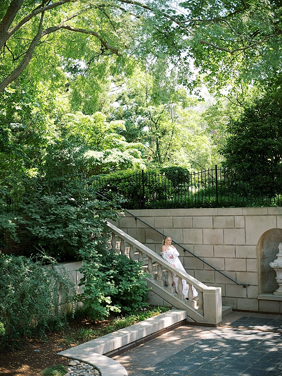 Bridal portrait of a bride in an off the shoulder wedding dress and veil standing on a stone staircase by garden greenery and fountain niche