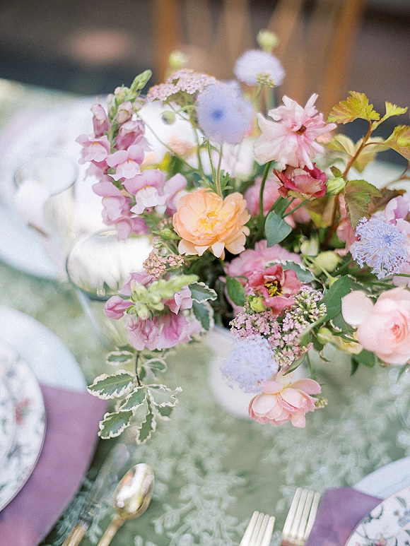 Wedding centerpiece with a pastel wedding centerpiece of mixed blooms and greenery on a white tablecloth with patterned plates and purple napkins