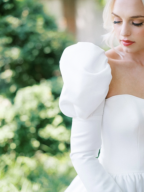Bridal portrait of a bride in a puff sleeve wedding dress with blonde updo and long lashes, set against soft garden greenery bokeh