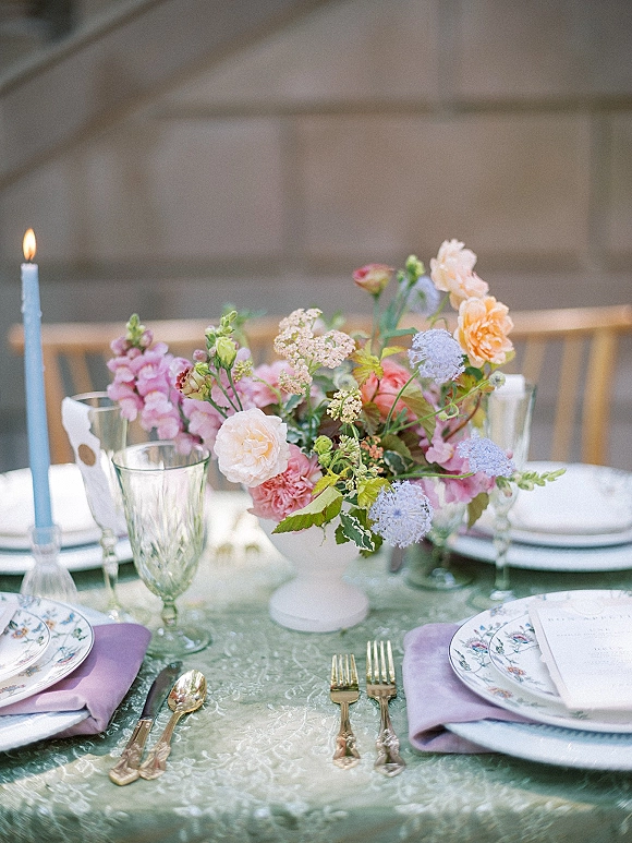 Reception tablescape with a wedding table centerpiece of pastel flowers in a compote vase, light blue taper candle, gold flatware, stone wall backdrop