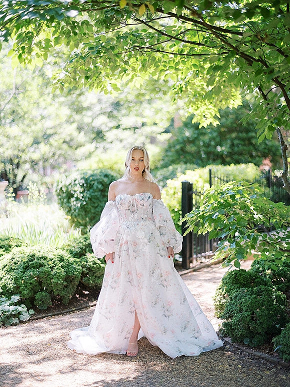 Bridal portrait of a bride in an off the shoulder wedding dress with sheer puff sleeves, standing on a sunlit garden path by an iron gate