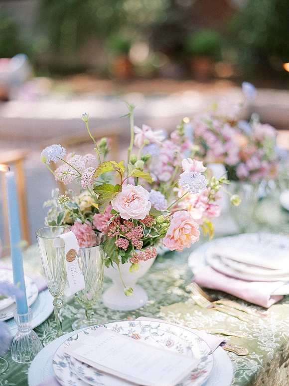 Reception tablescape with pastel floral centerpiece in white urn, blue taper candles, patterned china, pink napkins on a garden tablecloth outdoors