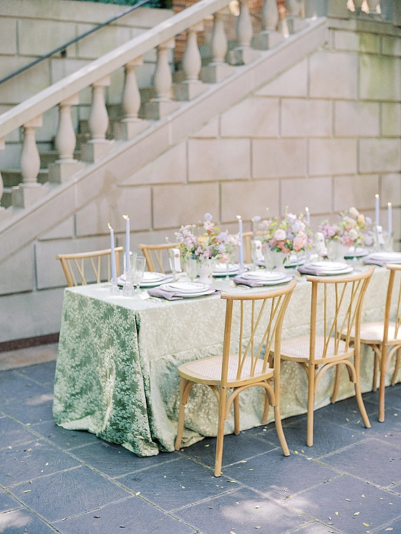 Reception tablescape with a green patterned tablecloth, pastel floral centerpieces and taper candles on a long table in a stone courtyard patio