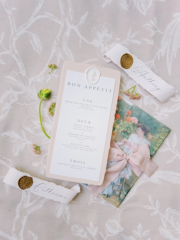 Wedding stationery flatlay featuring a wedding menu card, torn-edge place cards with gold wax seals, calligraphy, and ribbon on floral fabric