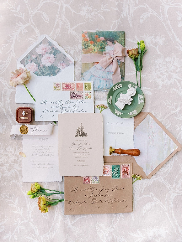 Invitation suite wedding invitation flat lay with deckled edge paper, calligraphy envelopes, wax seal, rings, and floral liner on patterned cloth