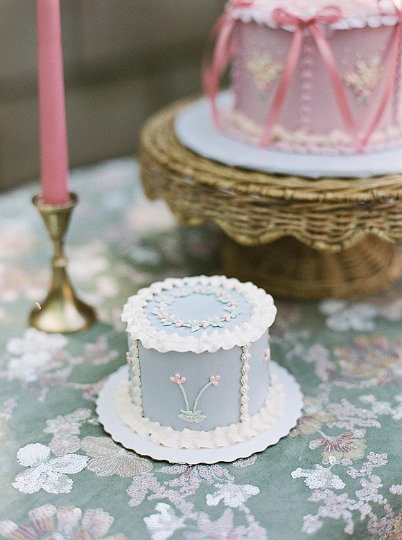 Wedding cake with piped ruffle icing and fondant flowers, tied with a pink ribbon on a stand beside a brass candlestick in greenery bokeh