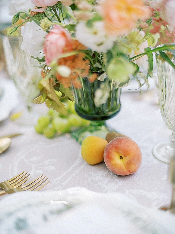 Reception tablescape with wedding table centerpiece in a green glass vase, stone fruit and grapes, gold flatware, and crystal stemware in soft light