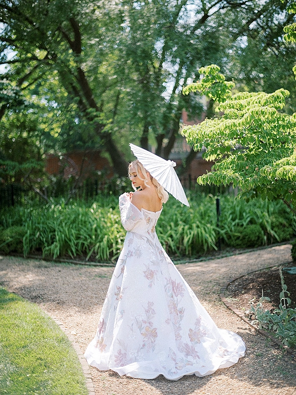 Bridal portrait of a bride in a pastel floral off-the-shoulder gown holding a white parasol on a gravel garden path among trees