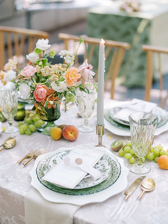 Reception tablescape with wedding table setting featuring a floral centerpiece, taper candle in brass holder, green goblets, patterned plates, and fruit on an outdoor patio