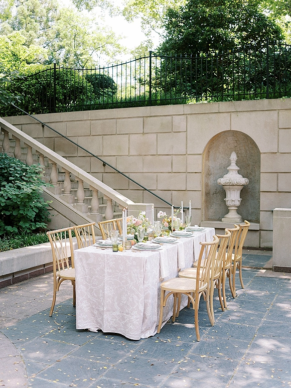 Reception tablescape with outdoor reception table on a stone patio, featuring a white patterned tablecloth, florals and taper candles
