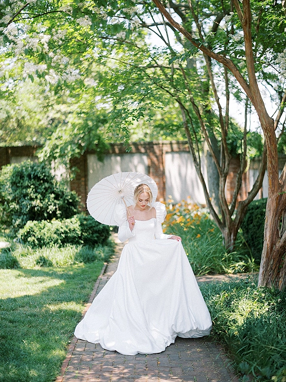 Bridal portrait of a bride with parasol in a puff sleeve white wedding dress walking along a brick garden path with greenery and shrubs