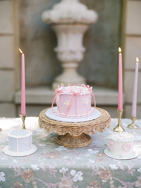 Wedding cake with pink frosted ribbon bows and piped buttercream on a wicker stand, surrounded by taper candles on a floral tablecloth outdoors