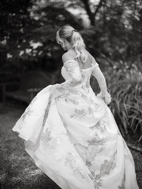 Bridal portrait in black and white of a bride walking away, holding her off the shoulder floral embroidered gown with lace gloves on a garden path