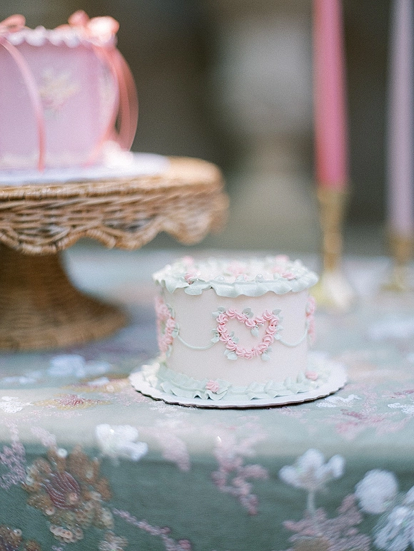 Wedding cake with pastel frosting and piped rosettes, heart detail on a wicker stand beside a taper candle on a floral tablecloth