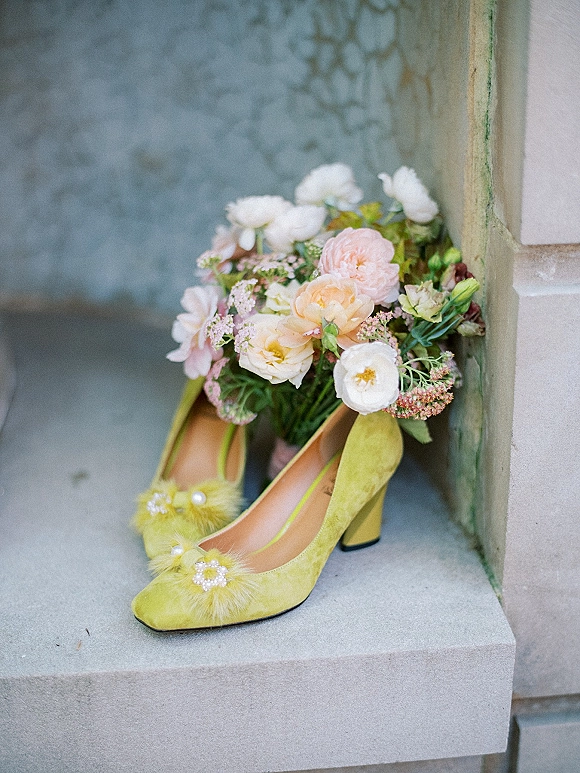 Wedding shoes in lime green heels with feather and crystal details beside a pastel bridal bouquet on stone steps by a stucco wall