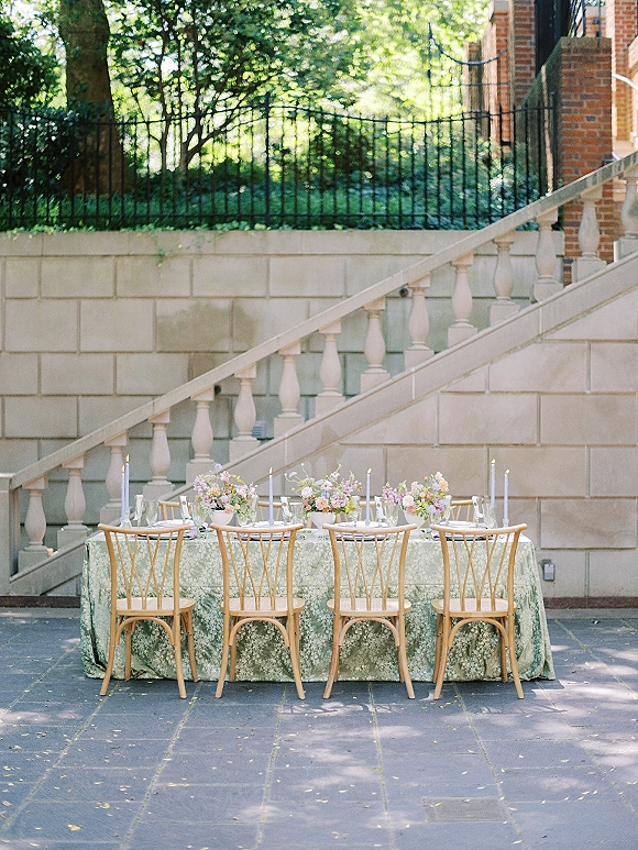Reception tablescape with an outdoor wedding dinner table set with floral centerpieces and taper candles on a stone patio courtyard by brick walls