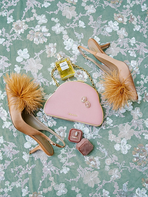 Bridal accessories flat lay with wedding details flat lay of pink clutch, nude slingback heels, feather clips, perfume, ring and earrings on green fabric