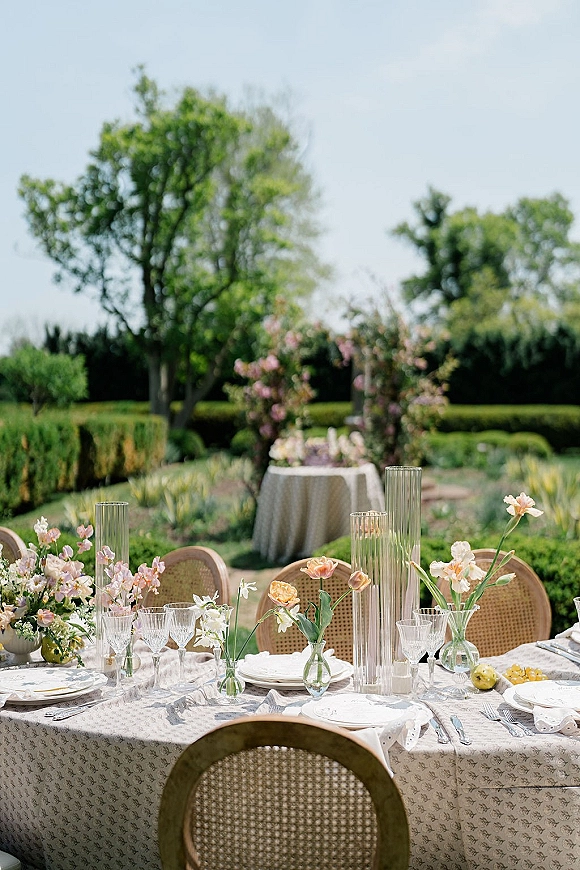 Reception tablescape with outdoor reception table settings, peach and blush flowers in tall cylinder vases, crystal glassware, rattan chairs in garden lawn