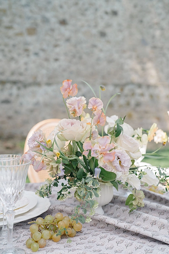 Reception tablescape with wedding floral centerpiece of pastel blooms in a white compote, textured goblets, grapes, set against a stone wall outdoors