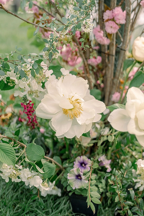 Wedding florals featuring white peony and soft pink blooms with greenery and flowering branches against a garden foliage backdrop