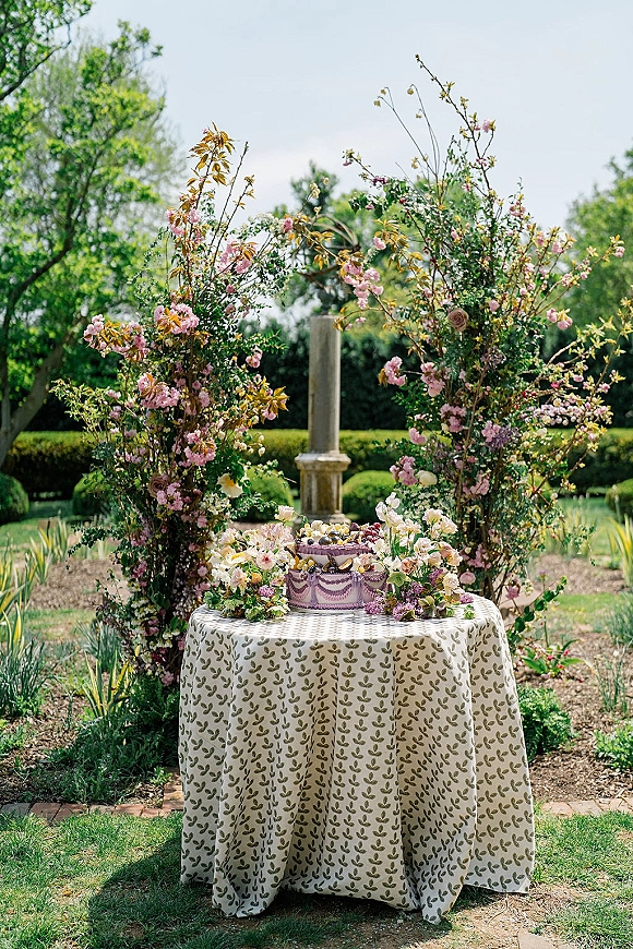 Wedding cake table with a single-tier buttercream cake, fruit garnish, blossom branches and greenery on a lawn garden backdrop