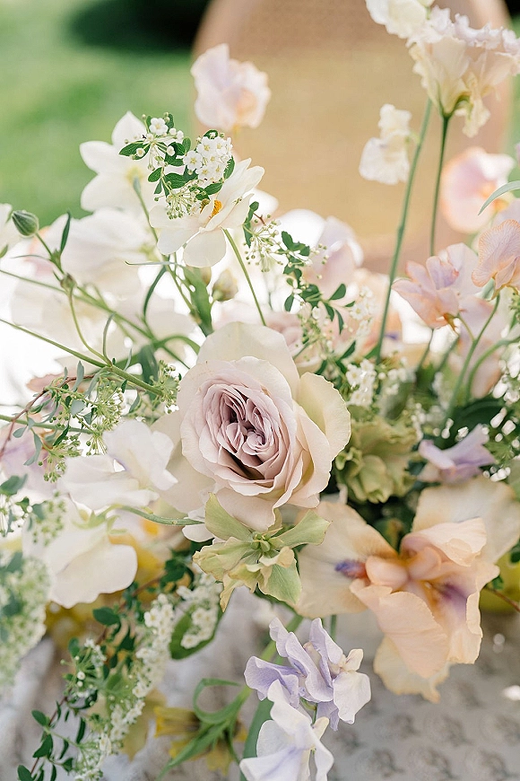 Wedding centerpiece of pastel wedding centerpiece roses, white flowers and sweet peas in a glass vase, set on an outdoor lawn with soft greenery bokeh