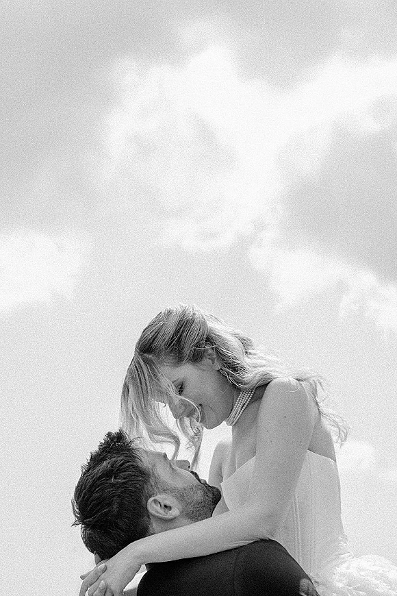 Couple portrait in a black and white wedding portrait style, groom holding bride in strapless dress with pearl choker, foreheads touching under a cloudy sky