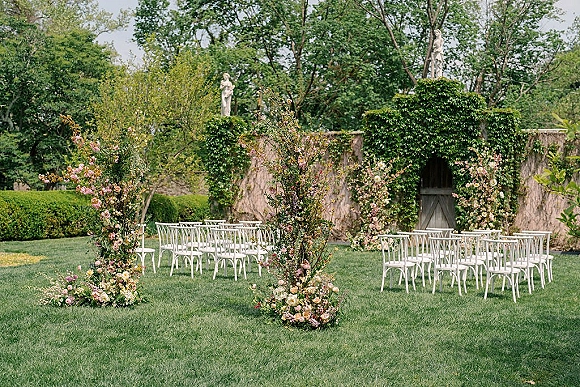 Ceremony setup for an outdoor wedding ceremony setup with a floral arch and white chairs on a lawn beside an ivy stone wall and gate