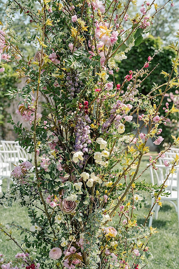 Wedding floral installation with flowering branches and pink blossoms framing an outdoor aisle beside white chairs on a garden lawn
