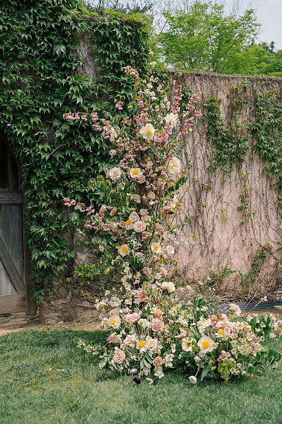 Wedding floral installation with pastel roses and climbing vines spilling across an ivy-covered stucco wall by a wooden gate outdoors