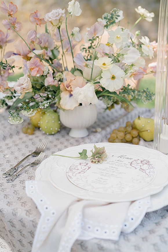 Reception tablescape with a wedding table centerpiece in a ceramic vase, menu card, taper candles, and fruit on an outdoor lawn