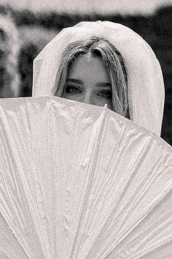 Bridal portrait in black and white of a bride with parasol, veil draped over her hair, peeking over the lace edge amid greenery