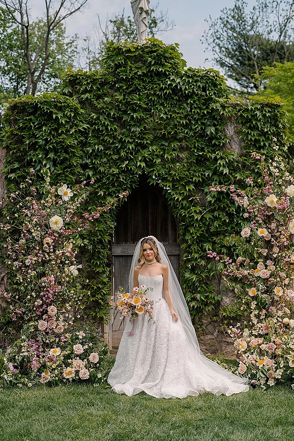 Bridal portrait of a bride holding bouquet in a strapless wedding dress, cathedral veil over her face, before a floral arch by an ivy wall