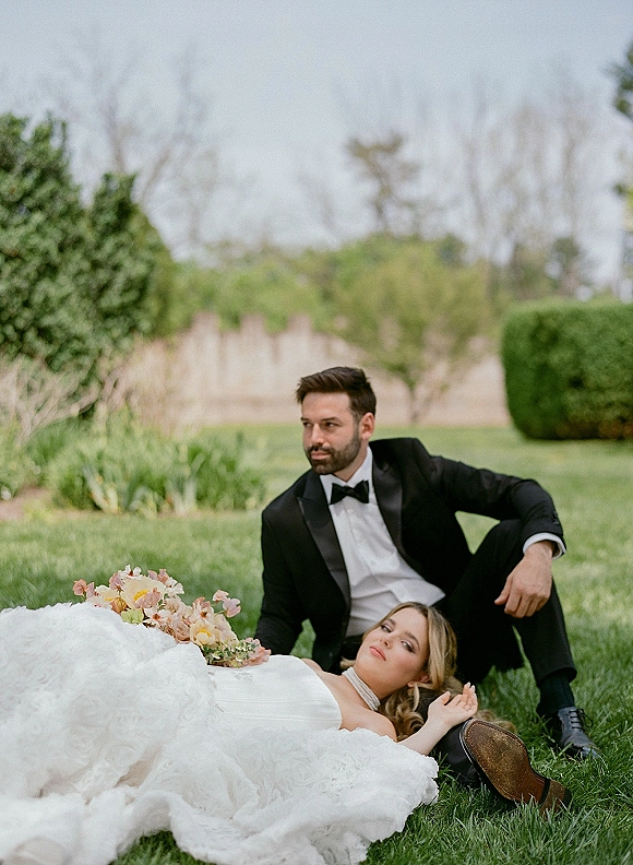 Couple portrait of bride in a strapless ruffled wedding dress reclining on groom in tuxedo with bow tie, holding pastel bouquet on a lawn