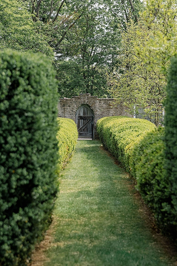 Garden venue entrance with an arched wooden door set in a stone wall, trimmed boxwood hedges framing a grass path under trees