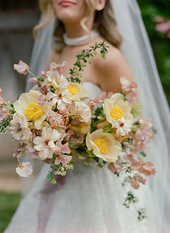 Bridal bouquet of pastel wedding bouquet blooms with greenery, held by a bride in a strapless dress and veil in a blurred garden backdrop