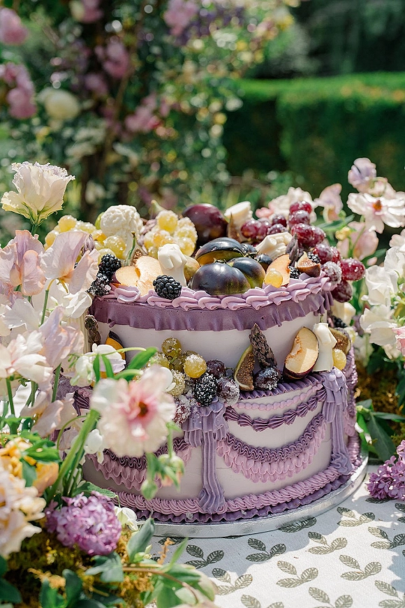 Wedding cake with lavender frosting ruffles, topped with figs and sugared berries, on a stand beside florals in garden greenery background