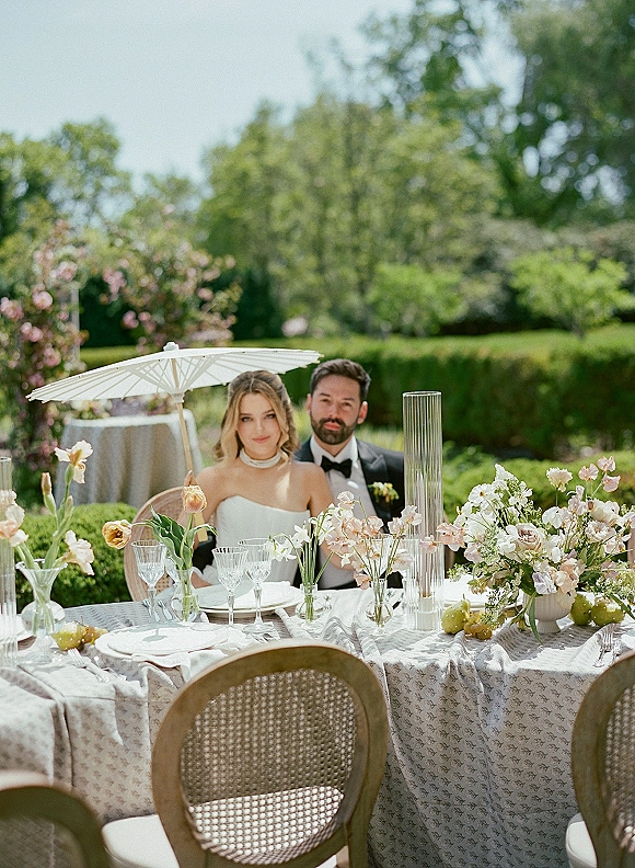 Couple portrait of bride in strapless gown and groom in tuxedo seated at a floral reception table with crystal goblets in a garden setting