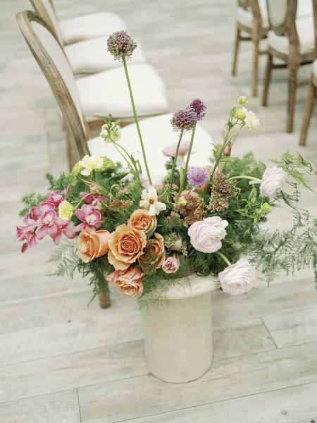 Wedding flower centerpiece with garden style blooms of roses, ranunculus, and allium with greenery in a vase on a wood floor by chairs