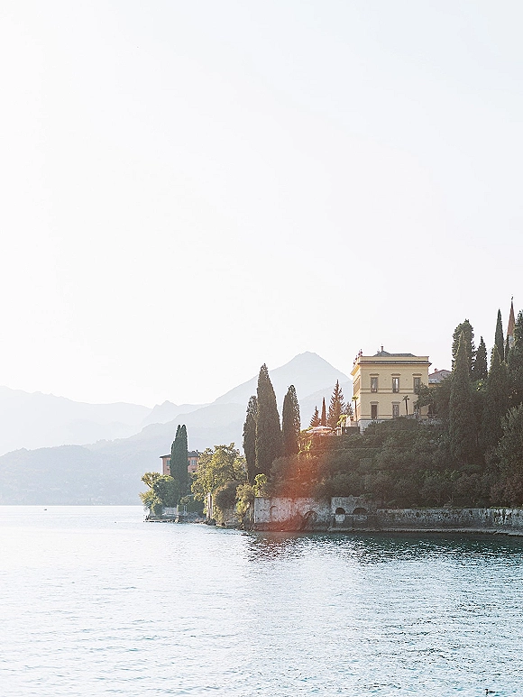 Wedding venue landscape with a lakefront wedding venue view of a hillside villa, cypress trees, and mountains along the shoreline under open sky