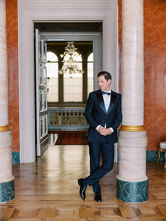 Groom portrait in a tuxedo with bow tie and cufflinks, standing in a grand doorway with marble columns and chandelier overhead