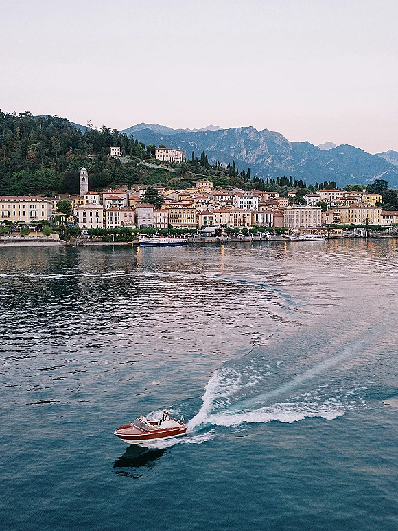 Wedding boat ride with newlyweds on speedboat in a wooden speedboat, gliding across a lake with mountains and a waterfront town behind