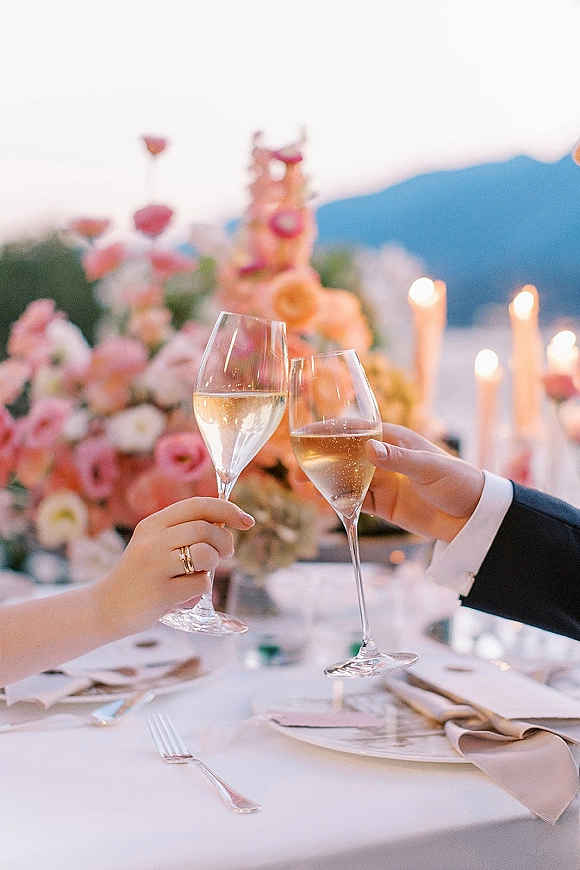 Wedding toast with champagne flutes raised beside a wedding ring, floral centerpiece and taper candles, with a mountain view behind