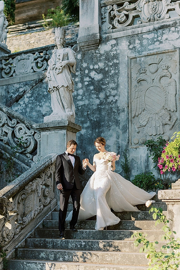 Couple portrait of bride and groom holding hands on a historic stone staircase, her strapless gown and soft pink bouquet beside a statue.